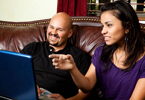A couple sitting on a couch and looking at a computer laptop screen.