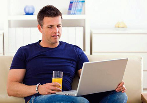 While sitting in his living room and drinking a glass of orange juice, a young male Veteran reviews the MyHealtheVet website on a laptop.