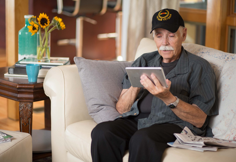 A Veteran sitting on a couch and using a handheld tablet to participate in a telehealth session.