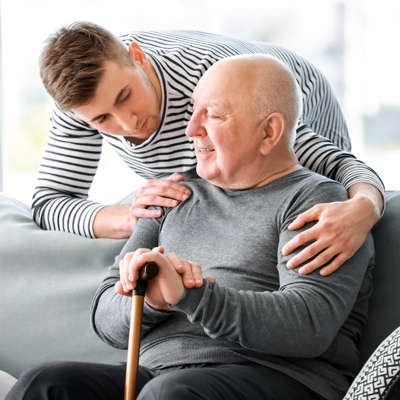A young man embraces his father sitting on a couch and holding a walking cane.