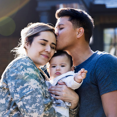A female Veteran holding her baby and being kissed by her partner.